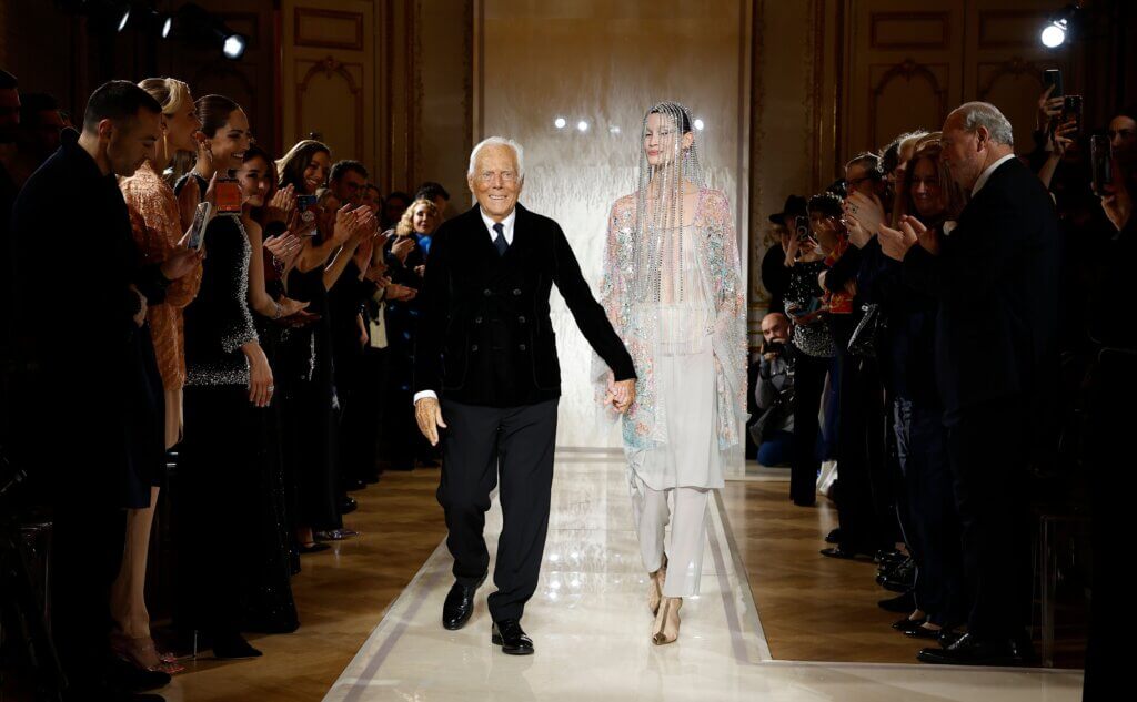 Giorgio Armani smiling and walking on a runway with an applauding audience in the background.