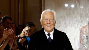Giorgio Armani smiling and walking on a runway with an applauding audience in the background.