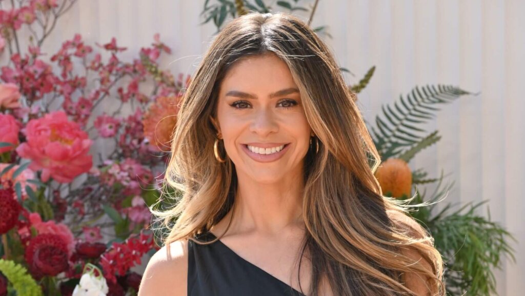 Smiling woman with long highlighted hair wearing gold hoop earrings and a black top, with pink flowers in the background.