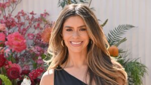Smiling woman with long highlighted hair wearing gold hoop earrings and a black top, with pink flowers in the background.