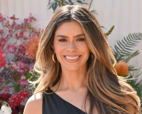Smiling woman with long highlighted hair wearing gold hoop earrings and a black top, with pink flowers in the background.