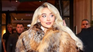 Blonde woman wearing a large fur coat looking to the side with people in the background