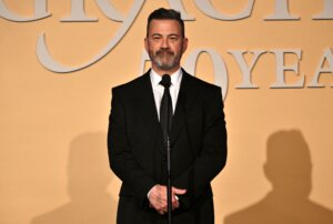Jimmy Kimmel standing at a microphone in a black suit and tie against a beige backdrop with partial text.