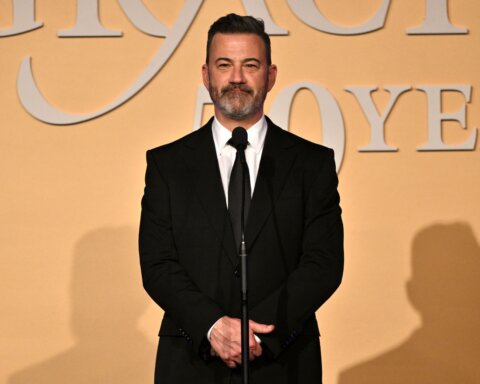 Jimmy Kimmel standing at a microphone in a black suit and tie against a beige backdrop with partial text.