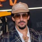 Actor and comedian Sebastian Maniscalco wearing a beige cap with "I WAS ON STAGE LAST NIGHT" text and sunglasses at a Netflix event