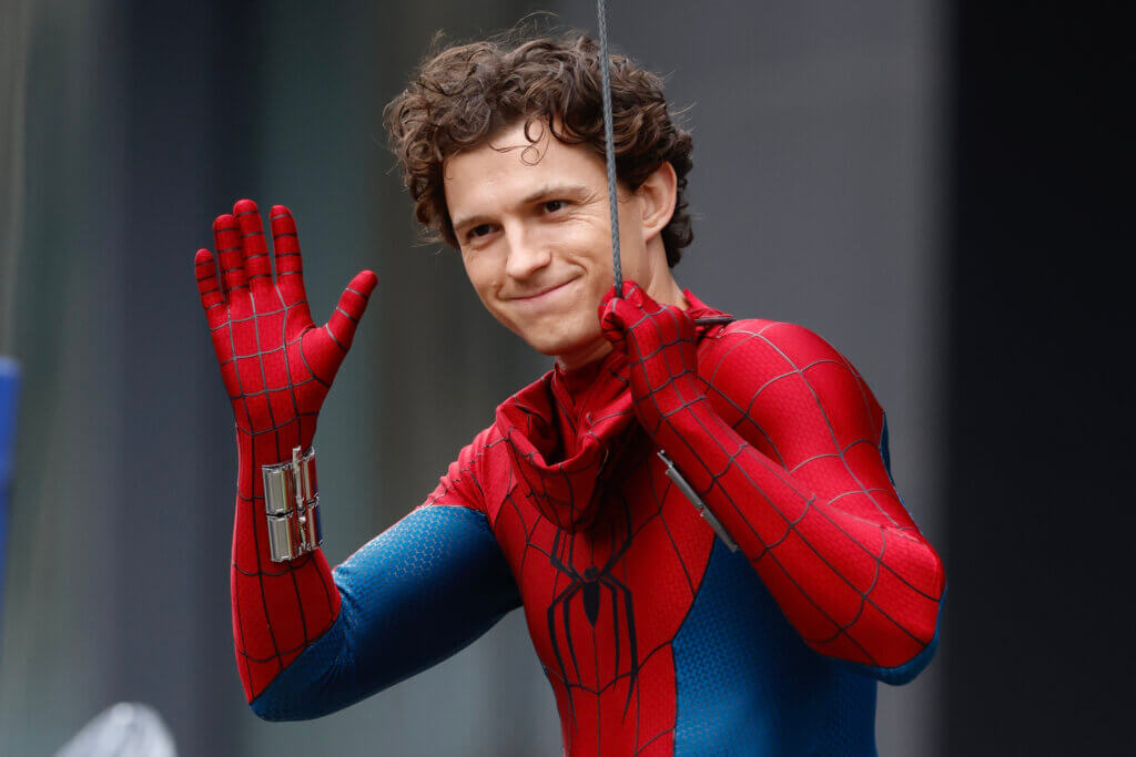 Tom Holland in Spider-Man costume waving while holding a web line