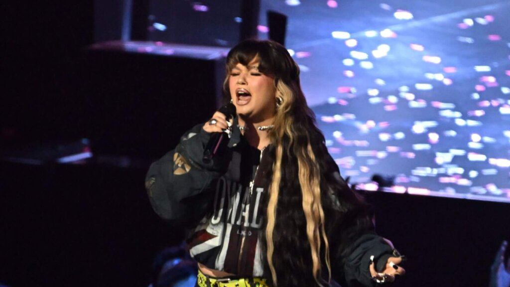 Singer with long black and blonde hair performing on stage with a microphone and colorful background lights