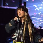 Singer with long black and blonde hair performing on stage with a microphone and colorful background lights