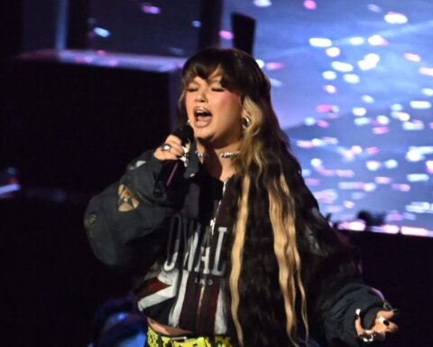 Singer with long black and blonde hair performing on stage with a microphone and colorful background lights