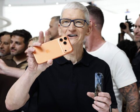 Tim Cook holding an orange iPhone 15 Pro Max at an Apple event with people and photographers in the background.