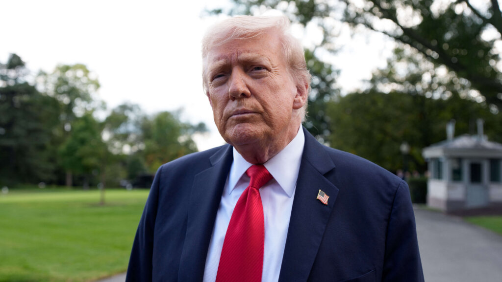 Donald Trump wearing a navy suit, red tie, and American flag pin outdoors with trees in the background.