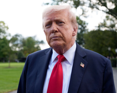 Donald Trump wearing a navy suit, red tie, and American flag pin outdoors with trees in the background.