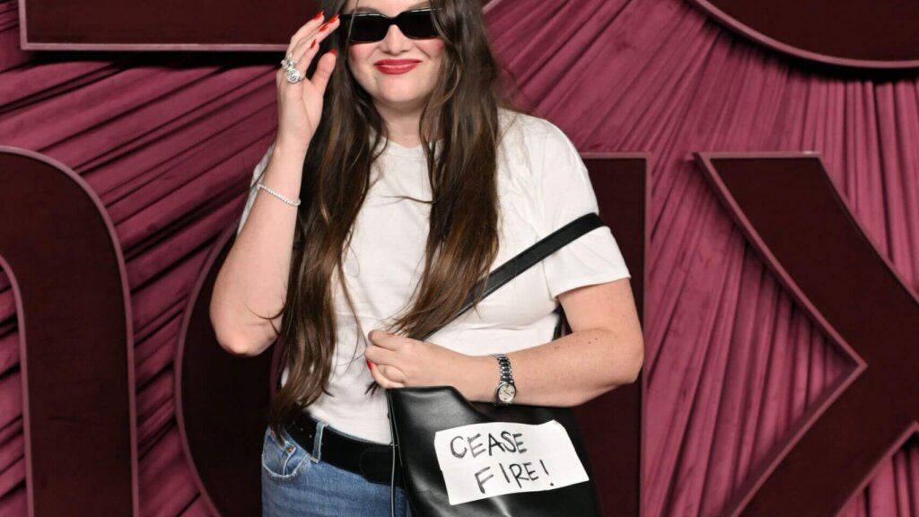 Woman wearing sunglasses, white t-shirt, and jeans holding a black bag with a "CEASE FIRE!" sign attached