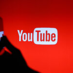 Silhouette of a hand holding a smartphone in front of a red screen displaying the YouTube logo.