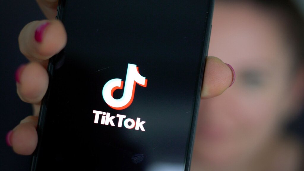 Hand holding smartphone displaying TikTok logo on screen with blurred background person