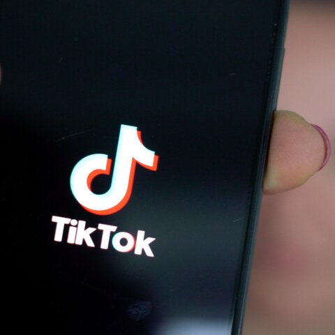 Hand holding smartphone displaying TikTok logo on screen with blurred background person
