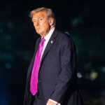 Donald Trump wearing a dark suit and bright pink tie walking outdoors at night with a "what's trending" logo in the corner.