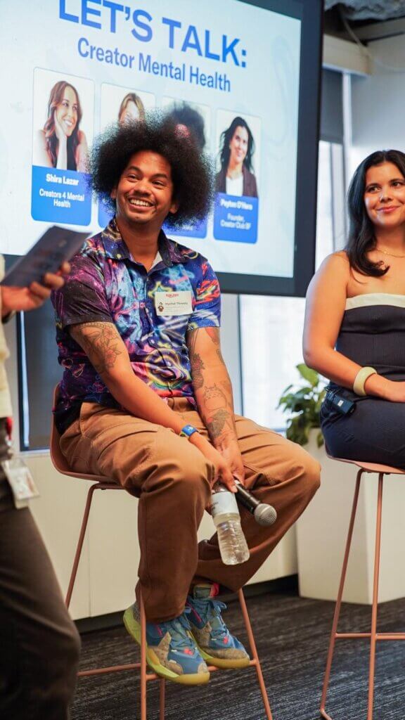 Mychal Theeist speaking at a panel with Shira Lazar and Peyton O'Hara shown on screen behind him