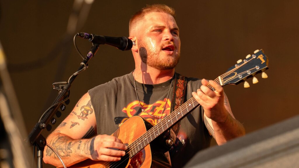 Male musician with tattoos playing a Gibson acoustic guitar and singing into a microphone on stage