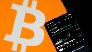 Smartphone displaying Bitcoin price at 111,843.60 USD with a rising graph against a large Bitcoin logo background.