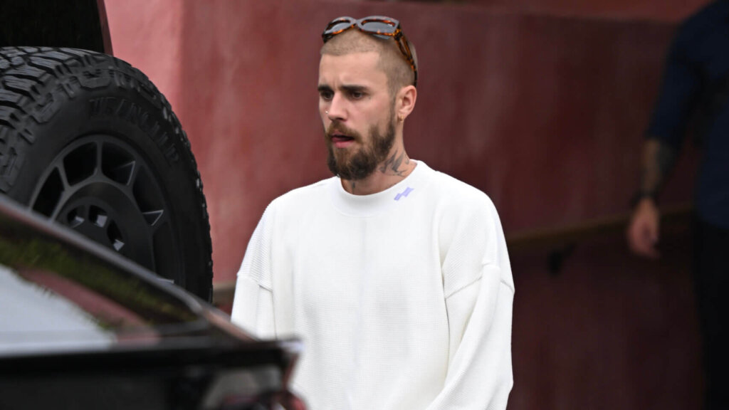 Man with beard and sunglasses on head wearing white sweater and pants standing near black Lamborghini car