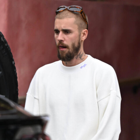 Man with beard and sunglasses on head wearing white sweater and pants standing near black Lamborghini car