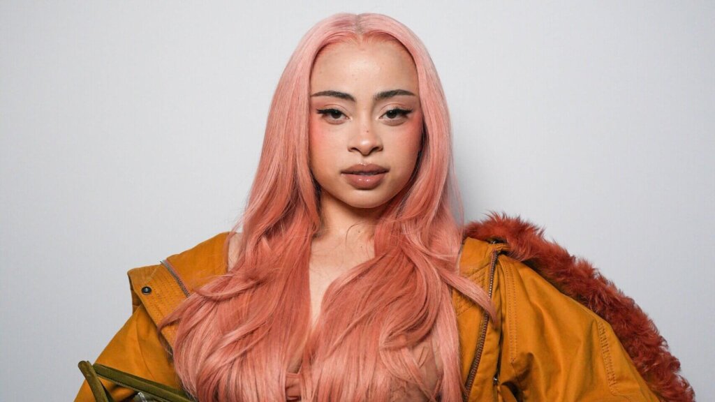 Woman with long pink hair wearing a sheer peach dress and oversized mustard jacket holding a green handbag against a gray background.