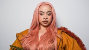 Woman with long pink hair wearing a sheer peach dress and oversized mustard jacket holding a green handbag against a gray background.