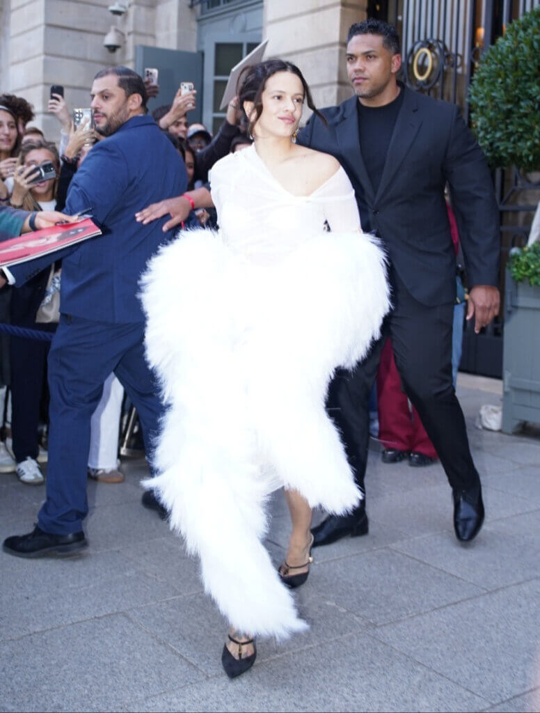 Rosalía in a white feathered dress walking with security and fans taking photos around her