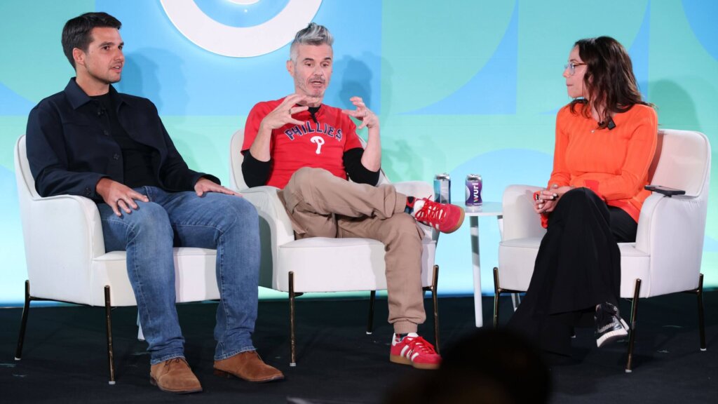 Three people seated on stage in a panel discussion, one wearing a red Phillies shirt and red sneakers.