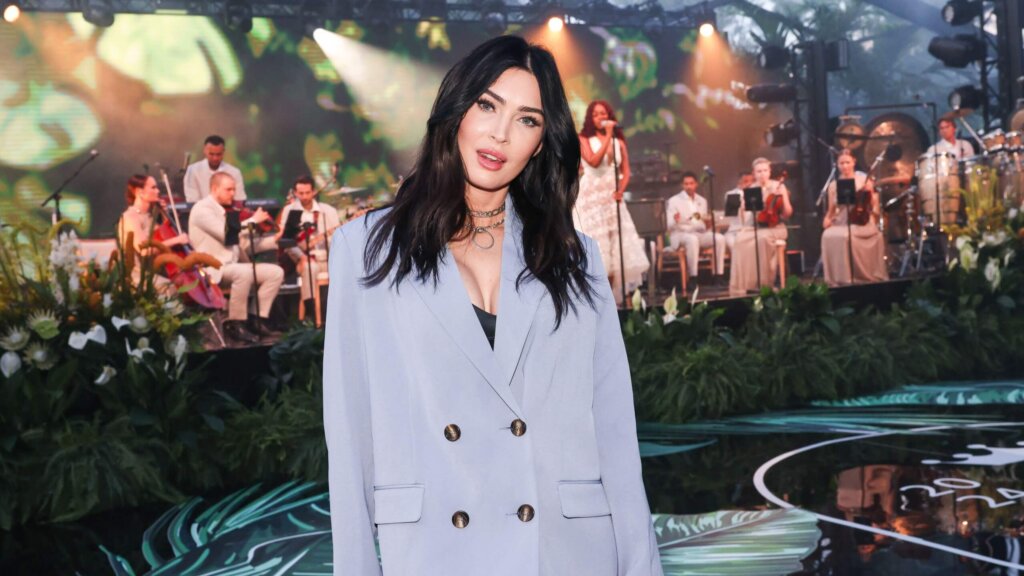 Woman in light blue suit holding red bag with live band performing on stage behind her