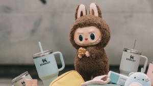 Stanley tumblers, colorful slippers, cosmetic products, and a brown bunny plush toy with "TREND > BRAND" graffiti in the background.