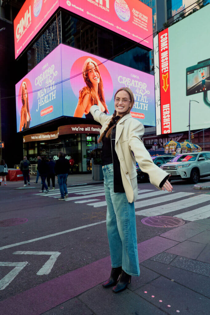 Woman smiling and posing in front of a Times Square billboard promoting mental health creator @haleyybaylee