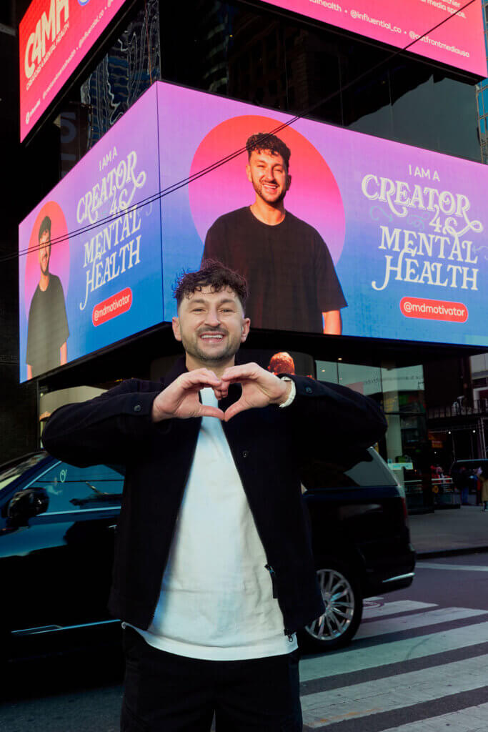 Man making a heart shape with hands in front of a digital billboard promoting mental health creator @mdmotivator