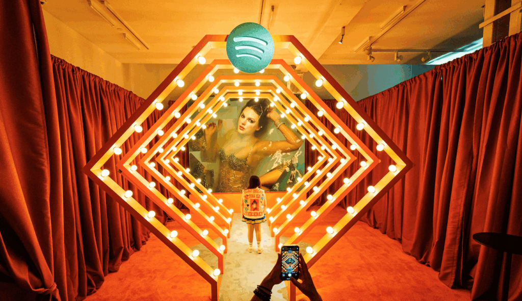 Person taking photo of another person standing in illuminated Spotify-themed art installation with red curtains.