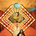 Person taking photo of another person standing in illuminated Spotify-themed art installation with red curtains.
