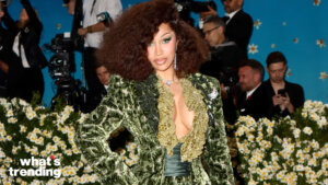 Cardi B wearing a green velvet ruffled dress posing at a floral-decorated event with photographers in the background