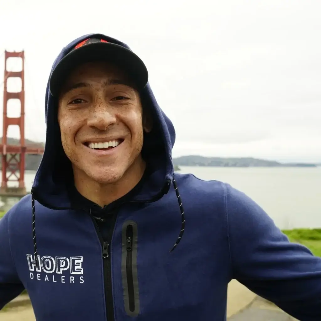 Smiling man wearing a navy hoodie with "Hope Dealers" text near the Golden Gate Bridge in San Francisco.