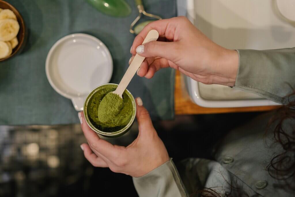 Hands holding a wooden scoop with green matcha powder over an open container on a table with sliced bananas nearby
