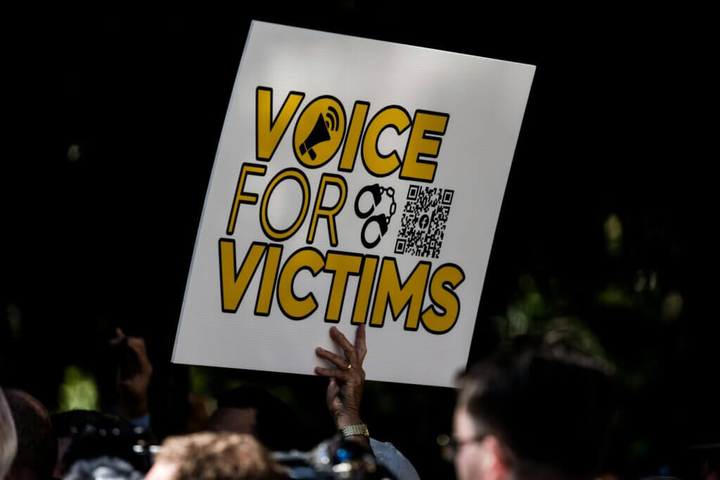 Hand holding a sign that reads "Voice for Victims" with a megaphone and handcuffs icon at a protest or rally.