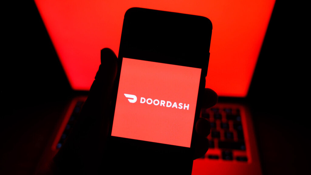 Hand holding smartphone displaying DoorDash logo with red laptop screen in background