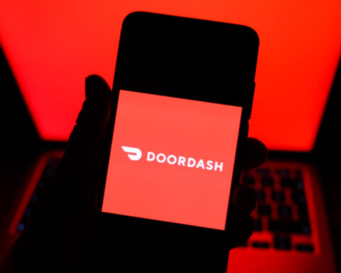 Hand holding smartphone displaying DoorDash logo with red laptop screen in background