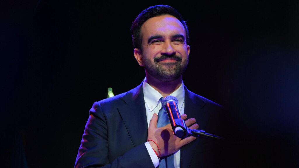 Man in suit and tie speaking at microphone with hand on chest against dark background