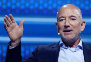 Jeff Bezos speaking with a headset microphone, gesturing with his left hand against a blue background.