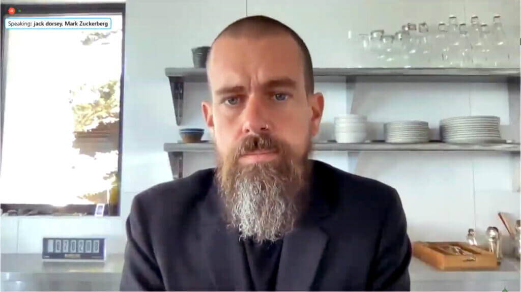 Jack Dorsey with a beard and nose ring speaking in a kitchen setting with shelves of dishes behind him.