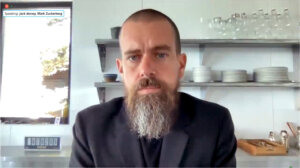 Jack Dorsey with a beard and nose ring speaking in a kitchen setting with shelves of dishes behind him.