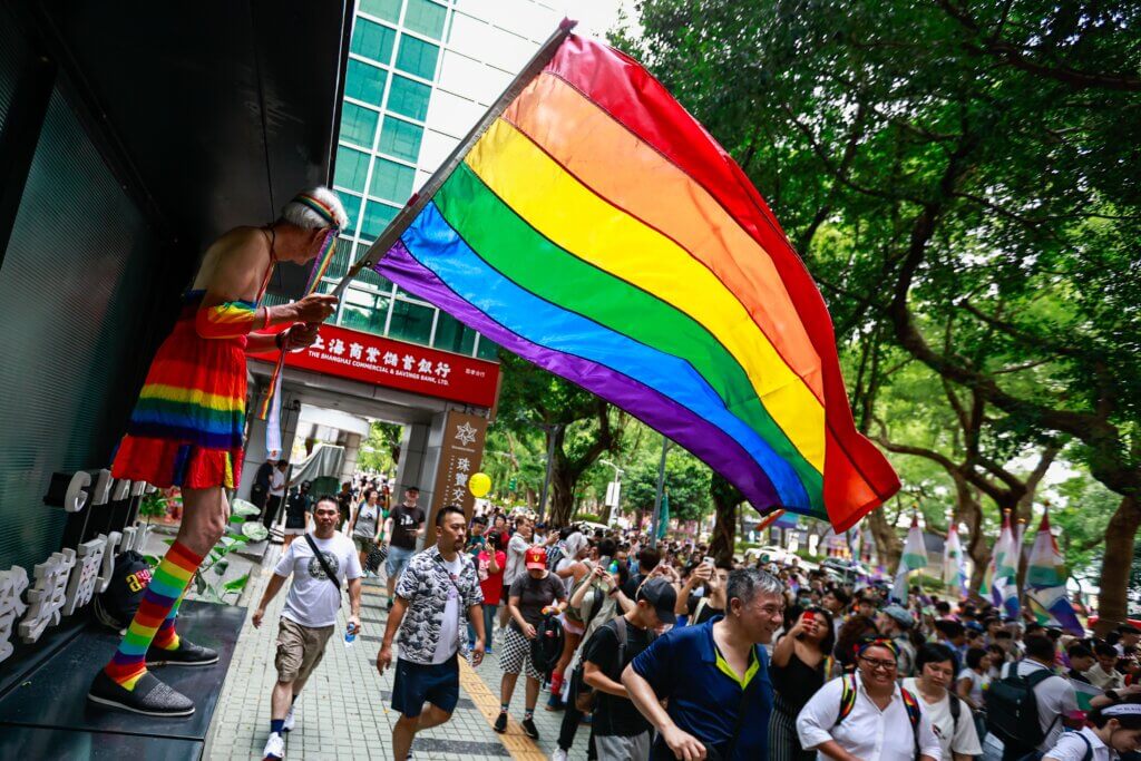 Person in rainbow outfit waving a large rainbow pride flag during a crowded outdoor event near The Shanghai Commercial & Savings Bank.