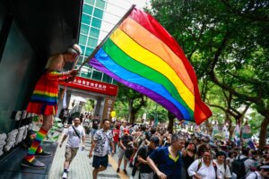 Person in rainbow outfit waving a large rainbow pride flag during a crowded outdoor event near The Shanghai Commercial & Savings Bank.