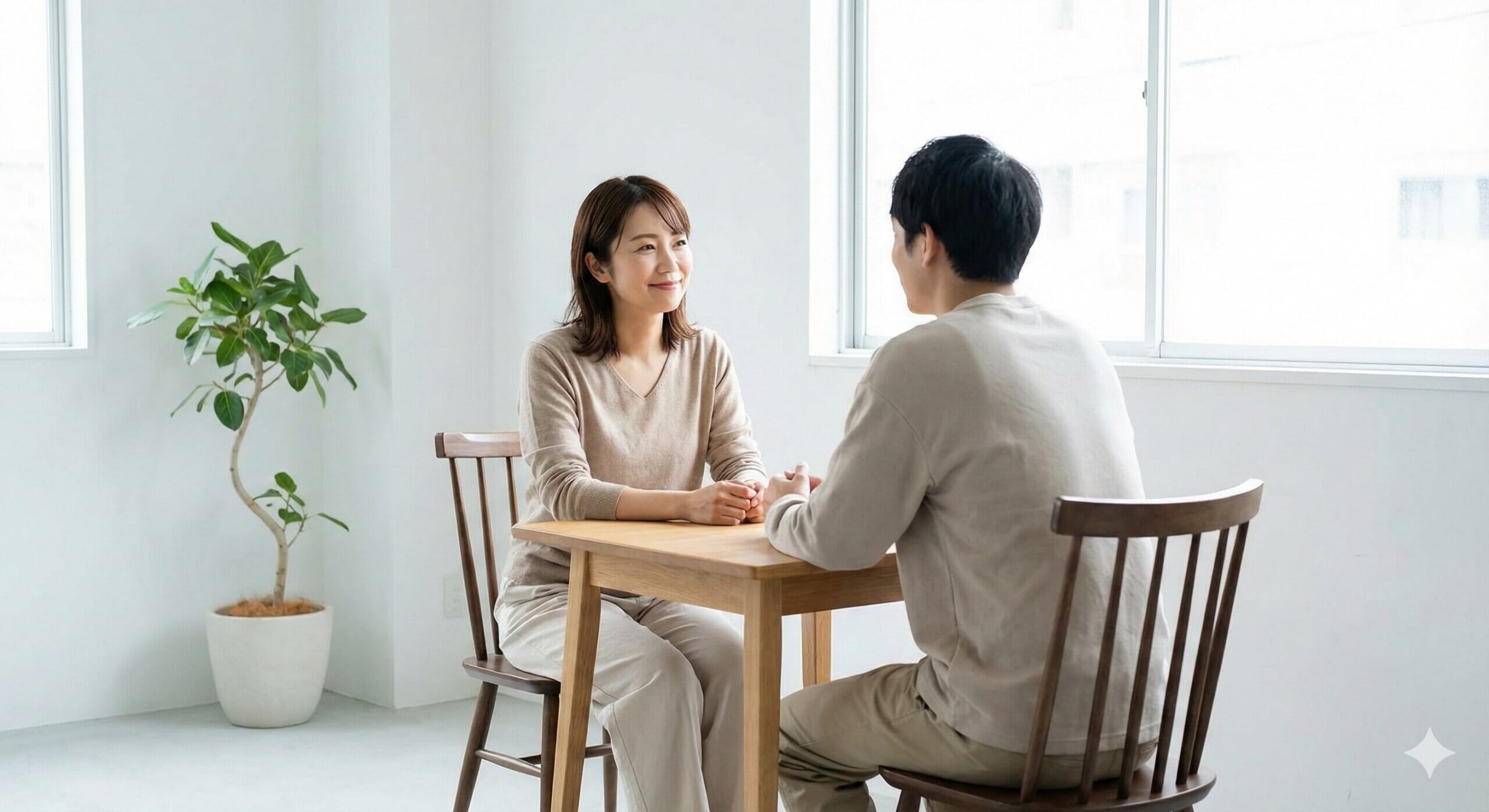 Two people sitting at a wooden table facing each other in a bright room with a potted plant nearby