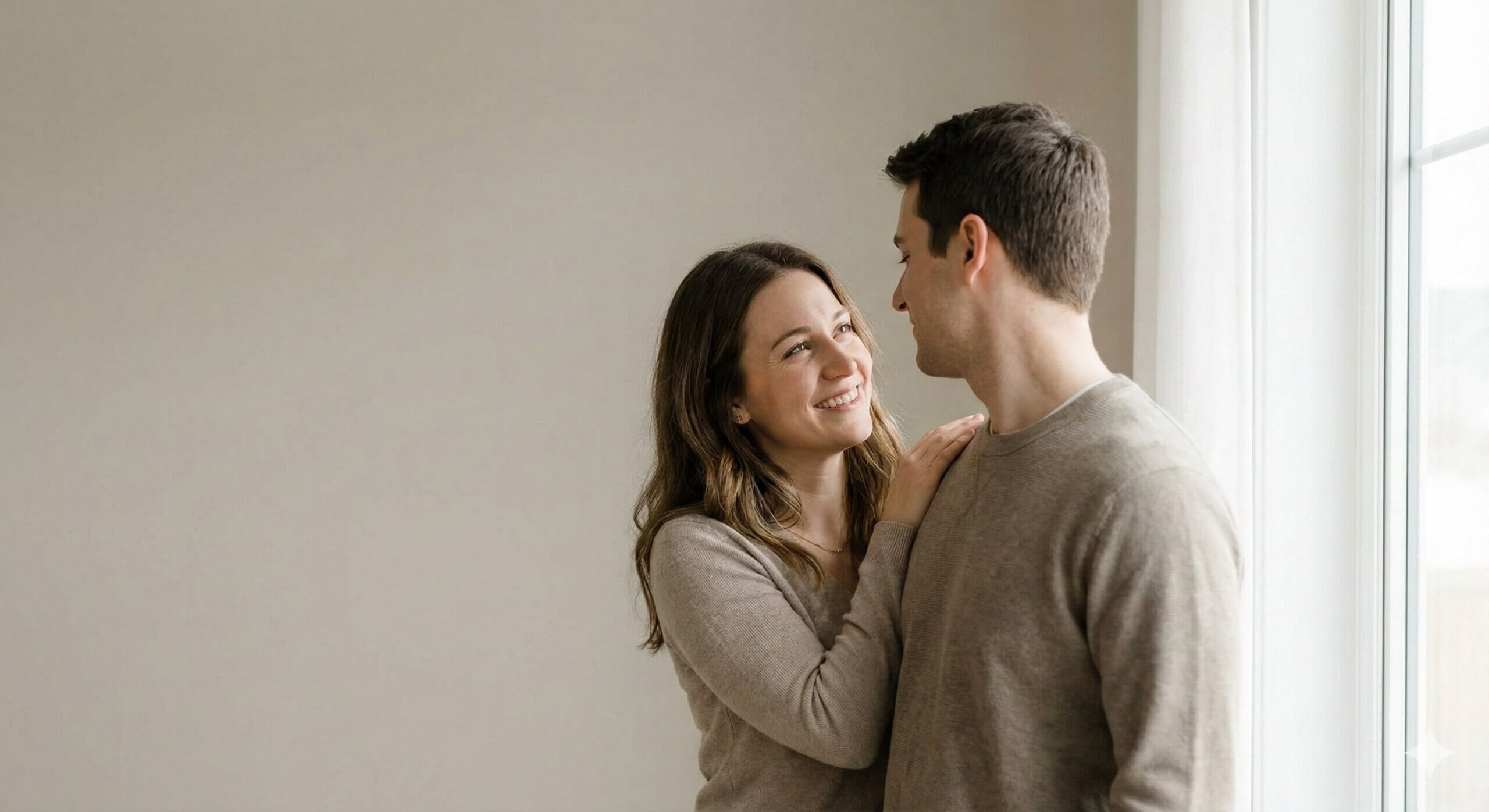 Smiling woman in beige sweater touching shoulder of man in beige sweater near window with white curtains